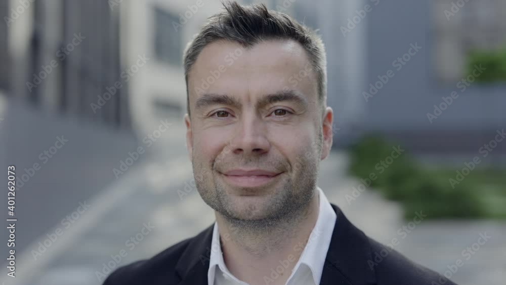 Close up view of positive mature enterpreneur looking to camera and smiling. Portrait of handsome man in 40s wearing suit posing outdoors near office buildings. Concept of success. Close up view of positive mature enterpreneur looking to camera and smiling. Portrait of handsome man in 40s wearing suit posing outdoors near office buildings. Concept of success.