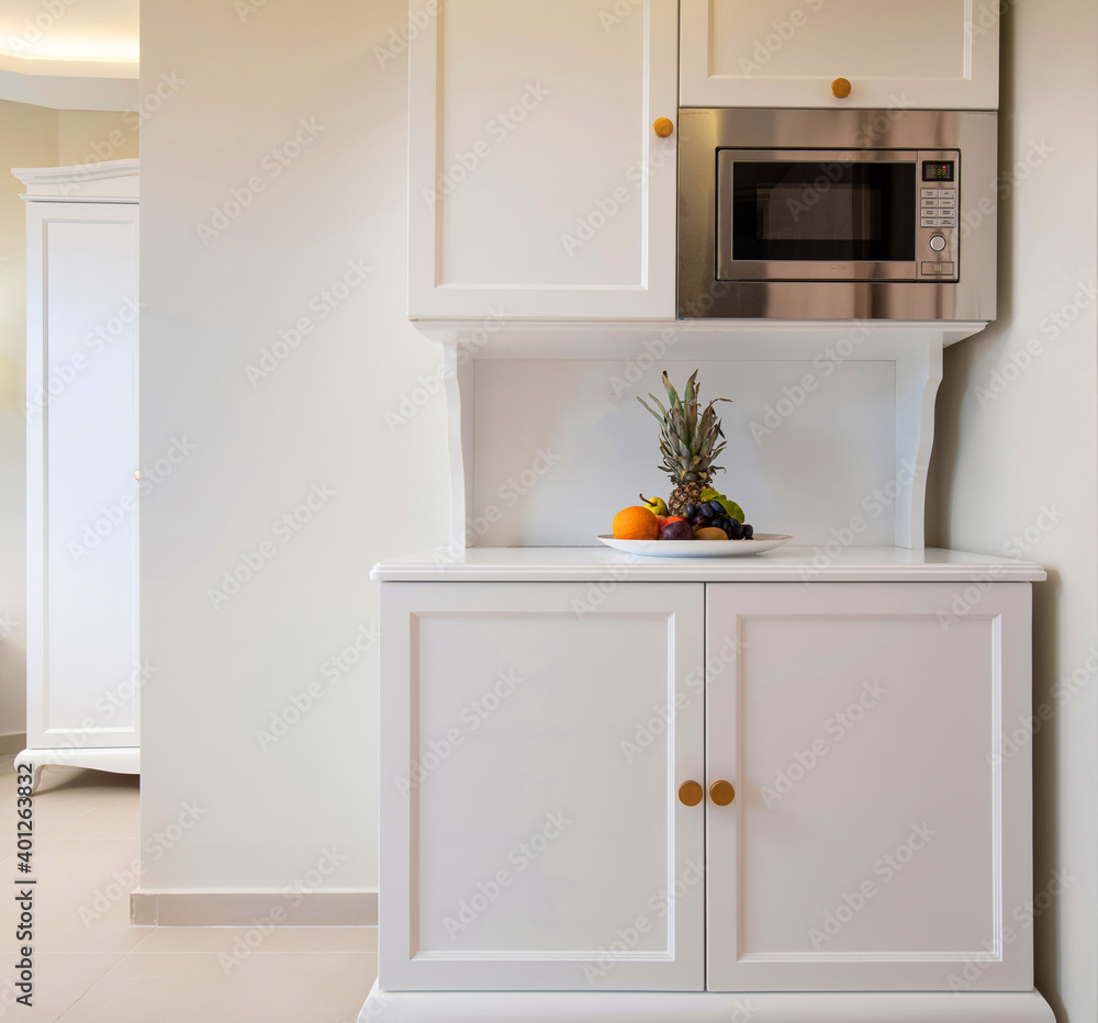 © GeorgeTsamakdas - Front view of mini kitchen cupboard module cabinet with microwave and fruits plate. Classic style interior of white room with empty copy space wall © GeorgeTsamakdas - Front view of mini kitchen cupboard module cabinet with microwave and fruits plate. Classic style interior of white room with empty copy space wall