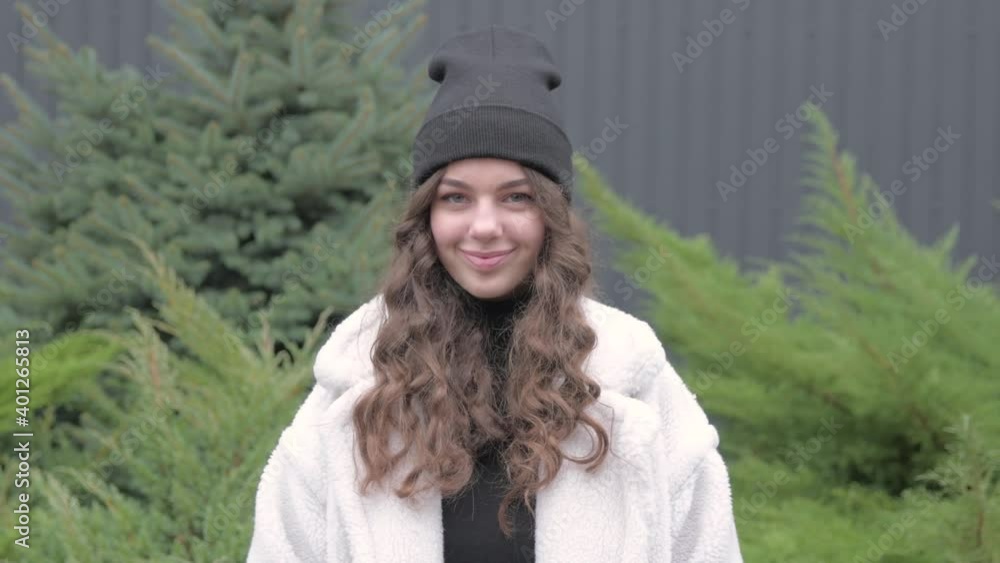 Looks and smile young adult european woman pretty face looking at camera posing alone standing at outdoor. front portrait. In warm clothes, jacket and hat in the yard.With flowing long hair. Looks and smile young adult european woman pretty face looking at camera posing alone standing at outdoor. front portrait. In warm clothes, jacket and hat in the yard.With flowing long hair.