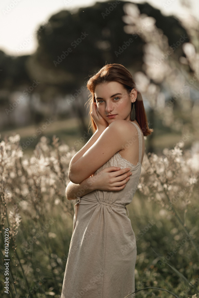 Medium shot portrait of young beautiful woman wearing a beige dress, posing in a field full of flowers and surrounded by trees. She is with her hand in her face. Medium shot portrait of young beautiful woman wearing a beige dress, posing in a field full of flowers and surrounded by trees. She is with her hand in her face.
