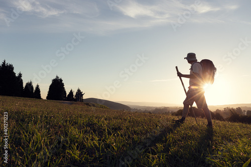 Side view of unrecognizable male pilgrim with rucksack and trekking stick strolling on green meadow under cloudy sky in sunshine in evening in back lit photo