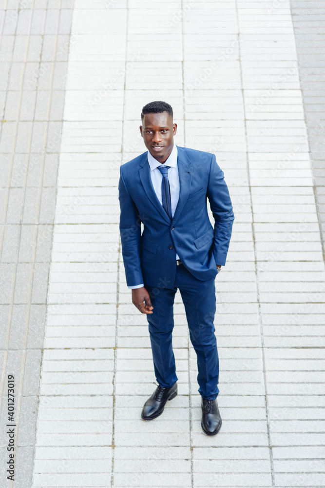 from above Confident adult African American male entrepreneur in elegant blue suit with tie looking at camera while standing against blurred urban background of modern city from above Confident adult African American male entrepreneur in elegant blue suit with tie looking at camera while standing against blurred urban background of modern city