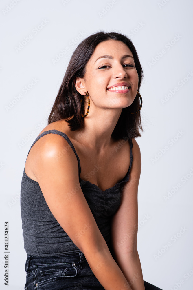 Full length young slim brunette in casual outfit sitting on stool against white background and looking at camera with happy smile Full length young slim brunette in casual outfit sitting on stool against white background and looking at camera with happy smile