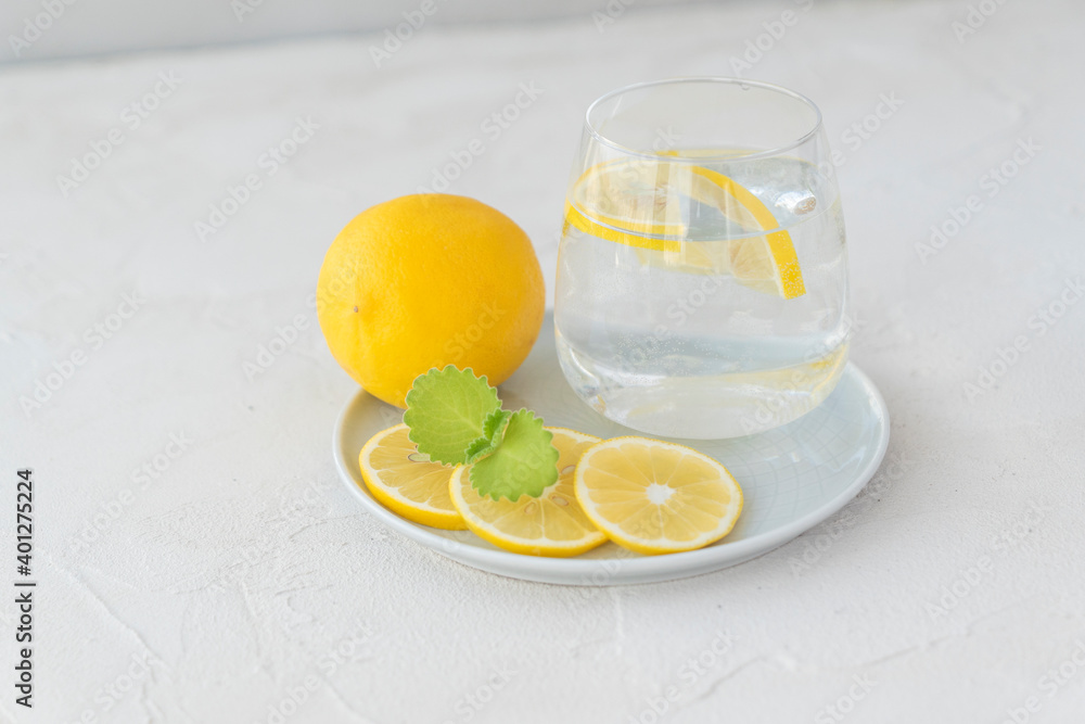 transparent glass with pure water, lemon slices, mint leaf , whole lemon lie on white plate on light background. Healthy vitamin drink transparent glass with pure water, lemon slices, mint leaf , whole lemon lie on white plate on light background. Healthy vitamin drink