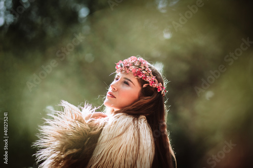 Side view of charming romantic young long haired female with pink floral wreath standing in forest looking away photo