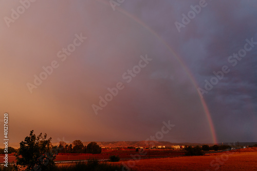 Scenery of vibrant rainbow shining on dark dramatic sky covered with thick heavy clouds in evening with vast agricultural fields in countryside