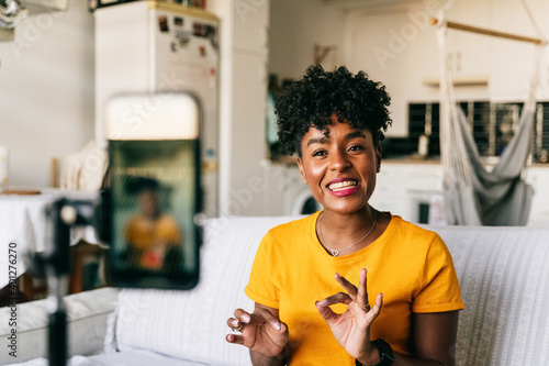 Cheerful young African American female blogger recording video on smartphone while preparing content for social networks at home
