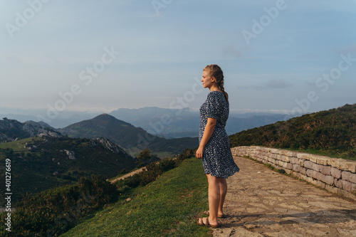 Side view full body young positive female impressed by amazing mountainous landscape standing on green hill and admiring views