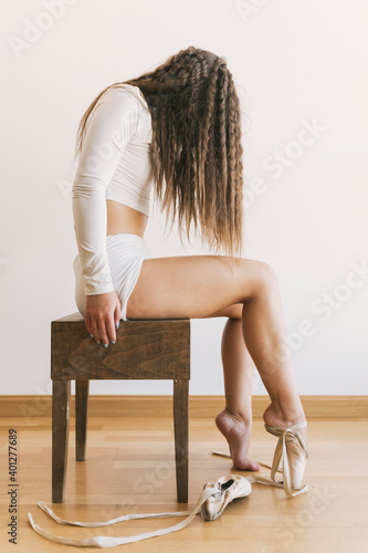 Side view of unrecognizable confident ballerina in pointe shoes sitting on stool in studio on white background
