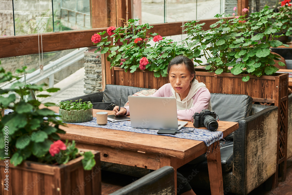 Concentrated Asian female journalist sitting at table with digital photo camera and using laptop while preparing new article Concentrated Asian female journalist sitting at table with digital photo camera and using laptop while preparing new article