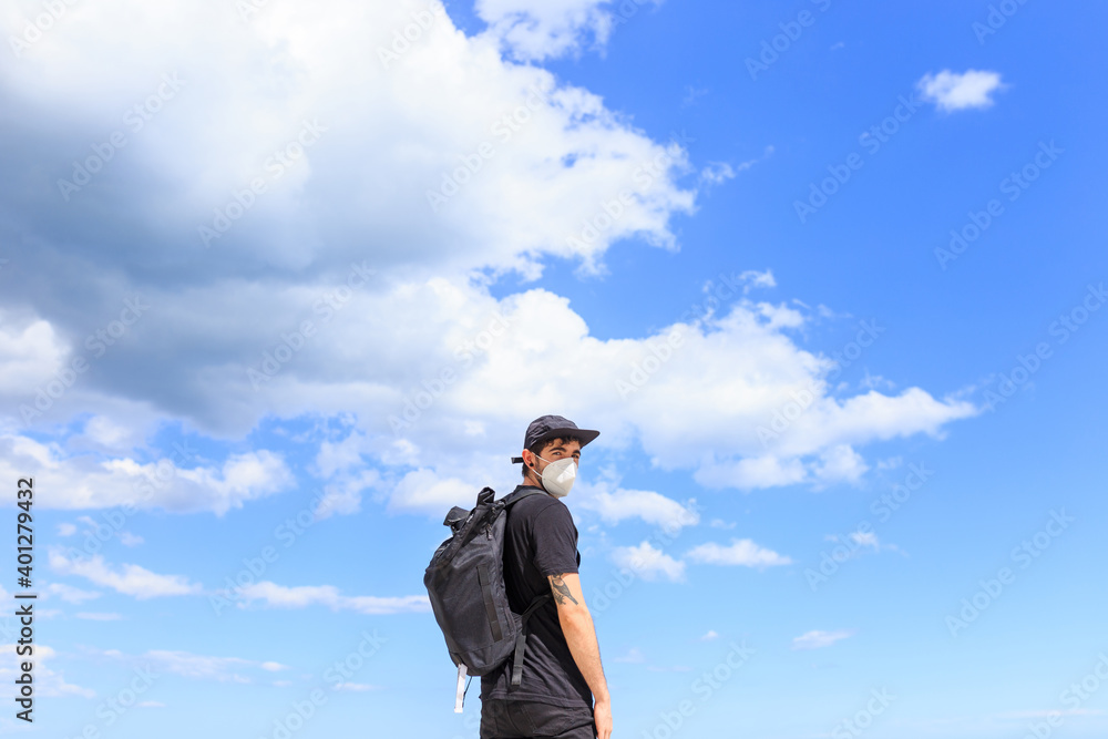 Low angle back view of male tourist in cap with backpack standing looking at camera under blue cloudy sky Low angle back view of male tourist in cap with backpack standing looking at camera under blue cloudy sky