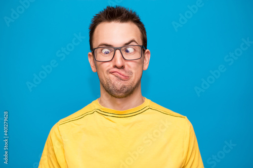 Portrait of young man wearing yellow shirt and trendy nerd glasses isolated over blue with confused face and funny expression © Svetlana