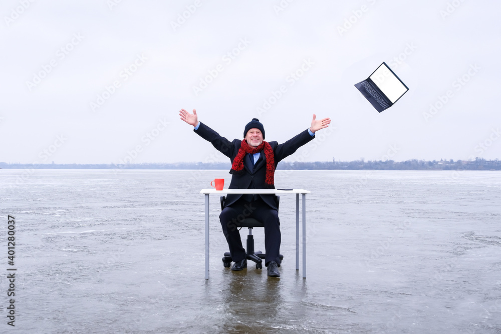 Obraz premium Senior smiling businessman in suit sits with outstretched hands at a table in the middle of a frozen lake in winter and throws away his laptop. Copy space for text on screen of device
