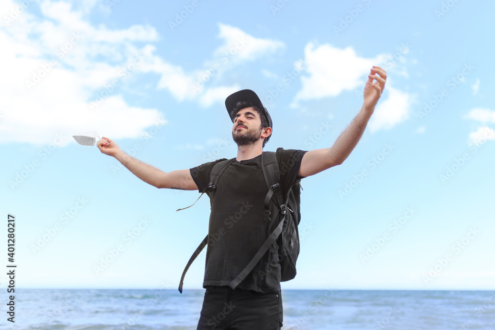 Low angle of smiling bearded male tourist in cap with backpack standing with raised arms and closed eyes behind ocean under blue cloudy sky Low angle of smiling bearded male tourist in cap with backpack standing with raised arms and closed eyes behind ocean under blue cloudy sky
