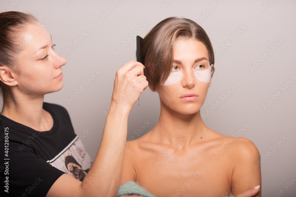 Female hairstylist with comb adjusting short hair of model standing with eye patches on violet background in studio Female hairstylist with comb adjusting short hair of model standing with eye patches on violet background in studio