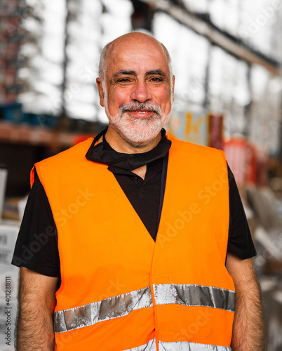 Serious adult male worker wearing orange high visibility vest standing near warehouse racks and looking at camera photo