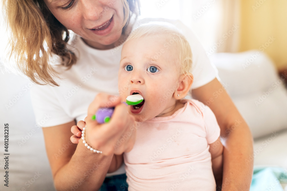 Crop smiling woman holding cute blond haired toddler and feeding with spoon in bright room on blurred background Crop smiling woman holding cute blond haired toddler and feeding with spoon in bright room on blurred background
