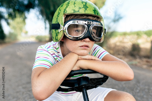 Crop dreamy boy in safety glasses and decorative helmet sitting leaned with hands on steering wheel on road while looking away photo