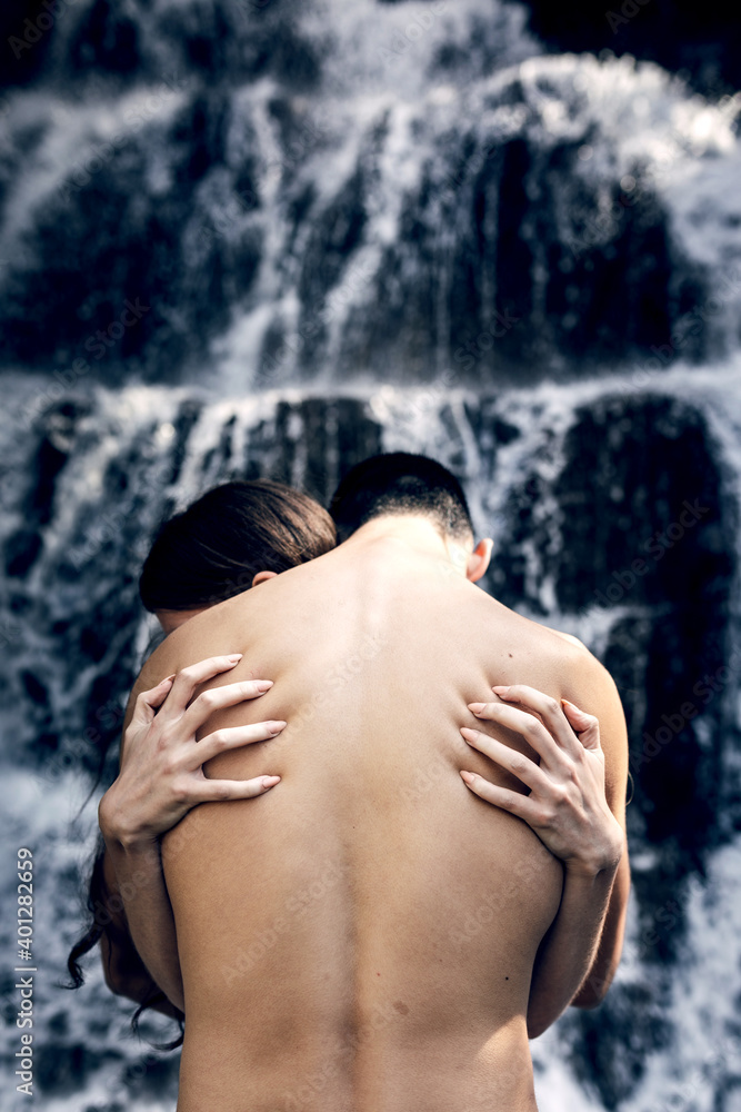 Back view of nude loving couple hugging tenderly while standing on background of waterfall in summer and enjoying nature together Back view of nude loving couple hugging tenderly while standing on background of waterfall in summer and enjoying nature together