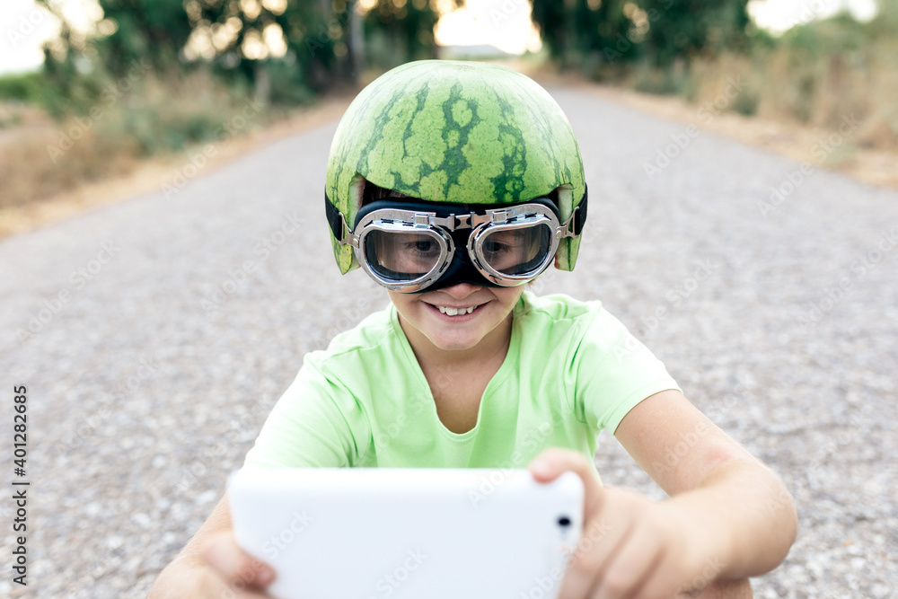 Low angle of cheerful kid in stylish wear and protective watermelon helmet taking selfie on cellphone while resting on skateboard on road Low angle of cheerful kid in stylish wear and protective watermelon helmet taking selfie on cellphone while resting on skateboard on road