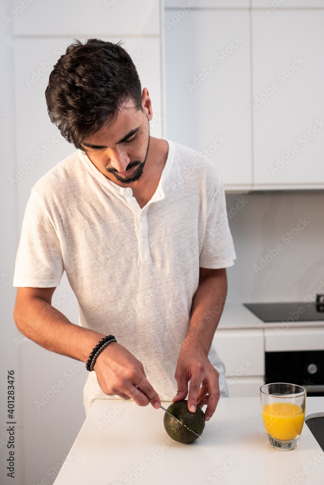 Serious male in domestic wear standing at counter in kitchen and cutting tasty avocado for breakfast in morning Serious male in domestic wear standing at counter in kitchen and cutting tasty avocado for breakfast in morning