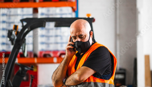 Serious adult male manager in uniform and protective mask standing with arms crossed near warehouse autoloader and having conversation on mobile phone photo