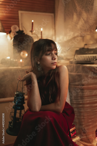 Peaceful female in red dress sitting in cozy room with old fashioned kerosene lantern while enjoying evening and looking away photo