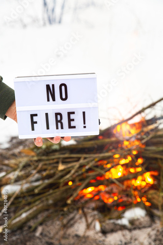 A sign with the text no fire in a man's hand over a flaming bonfire on a white background. The concept of nature protection, protection from forest fires, ecology. Open fire warning.