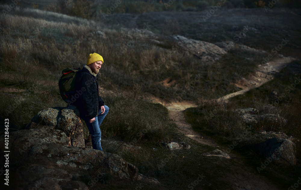 Obraz premium Side full-length portrait of a male traveller in yellow hat with a backpack on his back sitting on a rock on background of a walkway. Concept of people traveling in nature.