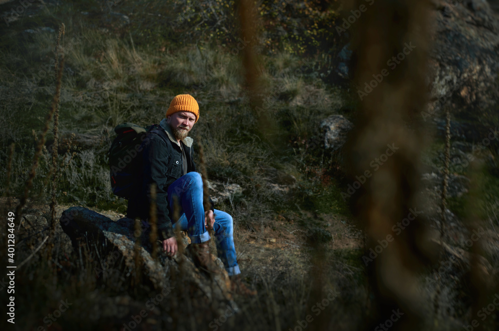 An adventurer man in an orange knitted woolen hat sat down to rest on a rock in the steppe forest and looks to the side. Concept of people traveling in nature.