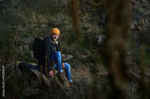 An adventurer man in an orange knitted woolen hat sat down to rest on a rock in the steppe forest and looks to the side. Concept of people traveling in nature.