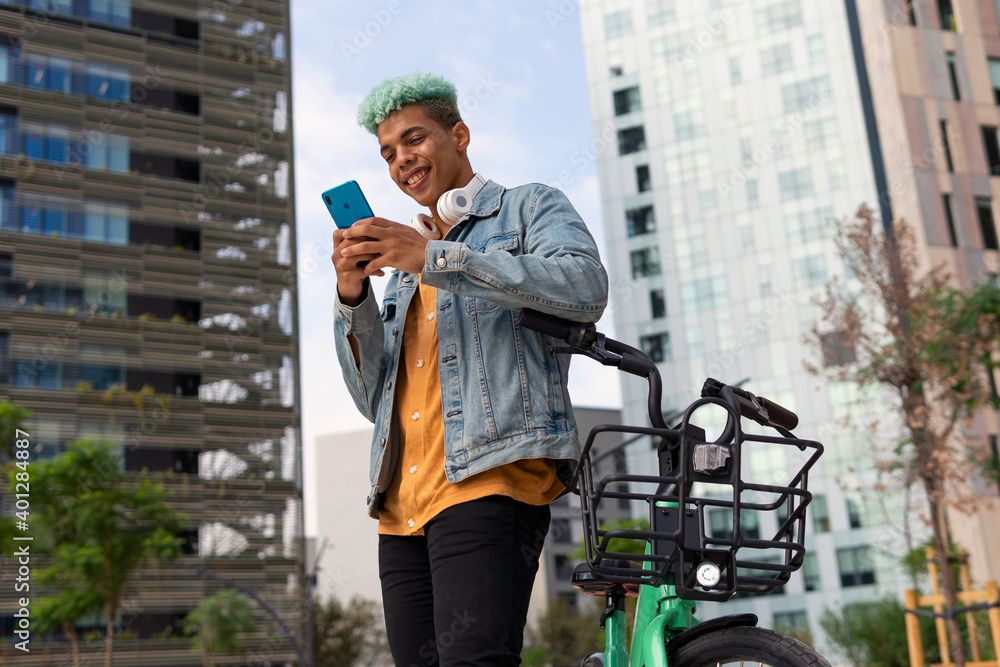 From below of positive handsome black male hipster with bright blue hair leaning on bike and chatting on social media via smartphone in city From below of positive handsome black male hipster with bright blue hair leaning on bike and chatting on social media via smartphone in city