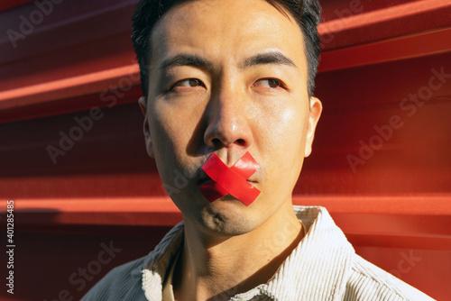 Crop pensive Asian male with red adhesive tape on lips standing against red metal fence and looking away thoughtfully photo