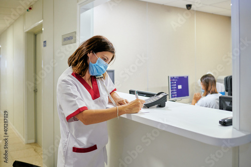 Concentrated female nurse wearing white medical suit and face mask taking notes in clinical record chart while standing near modern hospital reception photo