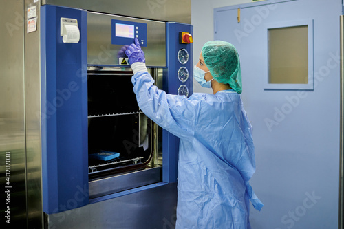 Side view female nurse wearing sterile protective uniform pushing buttons on modern sterilization machine while working in hospital