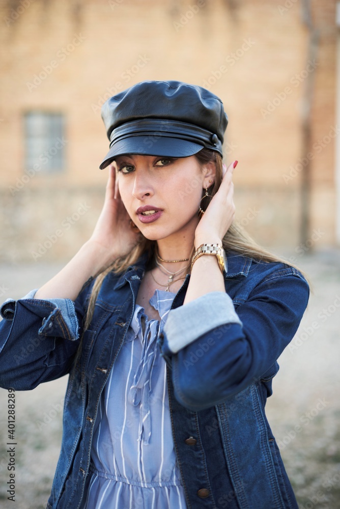 Side view of confident female wearing denim jacket and trendy leather cap standing on street and looking at camera Side view of confident female wearing denim jacket and trendy leather cap standing on street and looking at camera