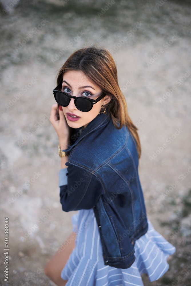 From above of cool young female wearing stylish casual clothes and sunglasses standing in nature and touching hair looking at camera From above of cool young female wearing stylish casual clothes and sunglasses standing in nature and touching hair looking at camera