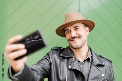 Cheerful glad bearded gallant male in hat taking photo in yard of green door
