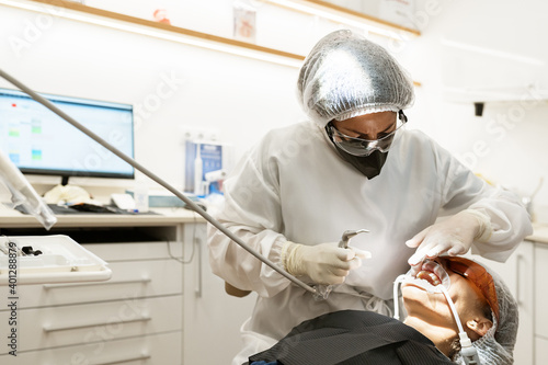 Professional dentist  in protective mask and goggles using drill machine while performing healing procedure to patient with saliva ejector and expander in mouth photo