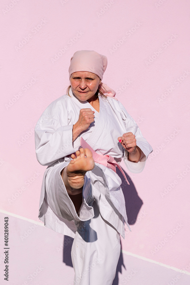 Determined mature woman in pink head cover and belt fighting karate in cancer battle concept in the street on pink wall looking at camera Determined mature woman in pink head cover and belt fighting karate in cancer battle concept in the street on pink wall looking at camera