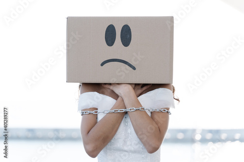 Anonymous female in white bridal dress with metal chain tying arms wearing cardboard box with unhappy smiley photo