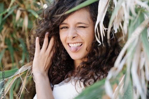 Flirty young female with curly hair looking at camera standing in foliage of verdant tree and showing tongue