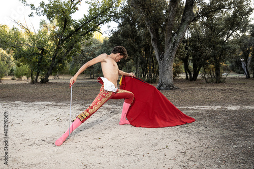 Focused slim male shirtless bullfighter in red pants holding cloak performing gracefully before corrida in nature