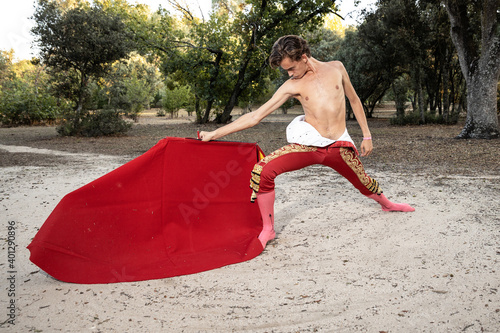 Focused slim male shirtless bullfighter in red pants holding cloak performing gracefully before corrida in nature photo