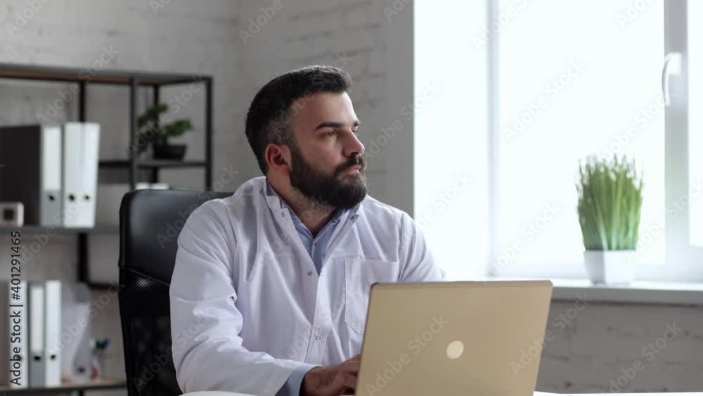 Handsome male doctor wearing white coat in the office consulting patients online using laptop. Family therapist texting messages on computer. Telemedicine, remote healthcare services concept Handsome male doctor wearing white coat in the office consulting patients online using laptop. Family therapist texting messages on computer. Telemedicine, remote healthcare services concept
