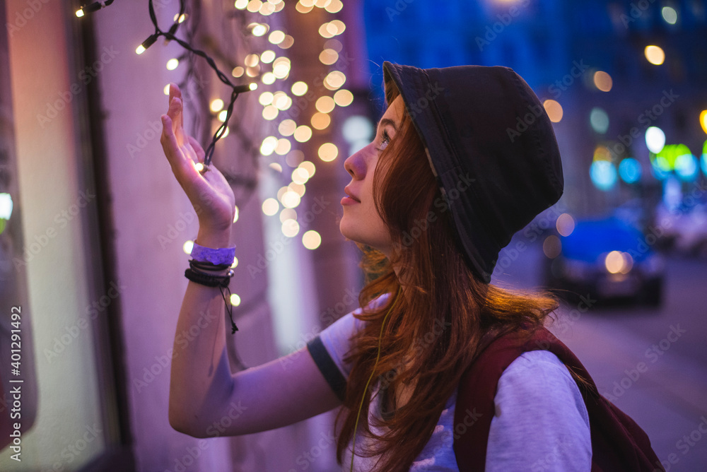 © Nacho Zaitsev/ADDICTIVE STOCK - Side view of stylish hipster millennial woman in hat and with backpack touching glowing garland while standing near building in evening city