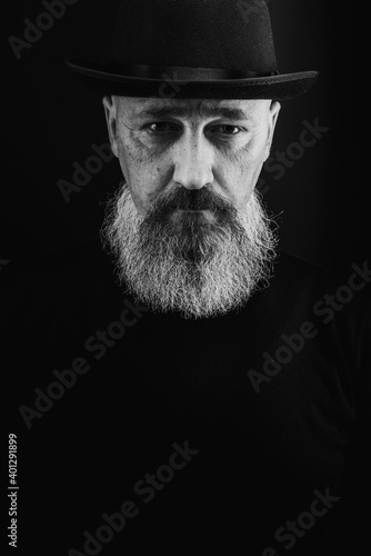 portrait of a young, handsome, emotional, serious man with a gray long beard and a black hat on his head, isolated on a dark background. There is a place for an inscription. black and white photo, low