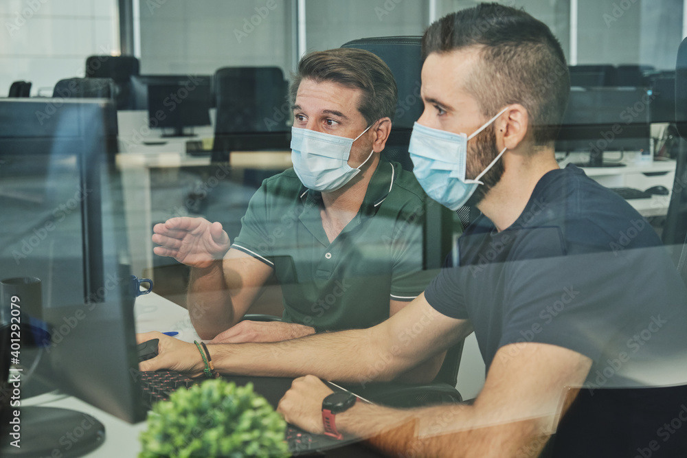 Male coworkers in medical masks sitting together using program for software during coronavirus pandemic