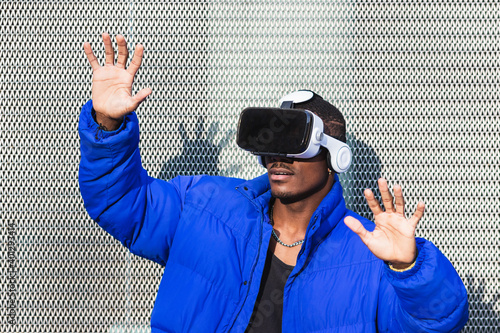 African American male standing on street near metal fence while exploring virtual reality in innovative goggles and touching air
