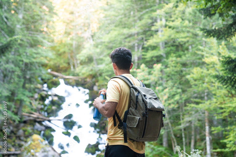 Back view of unrecognizable man traveler with backpack standing on slope with bottle drinking water while admiring amazing landscape of green forest with fast river Back view of unrecognizable man traveler with backpack standing on slope with bottle drinking water while admiring amazing landscape of green forest with fast river